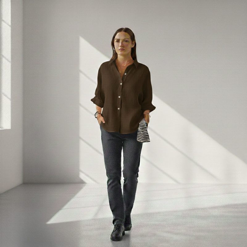 Woman in a brown shirt and jeans standing in a room with light streaming through a window.