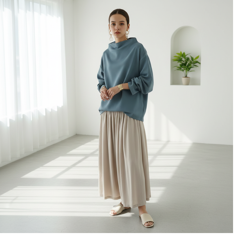 Woman wearing a blue sweater and beige skirt standing in a bright room with white walls and a plant.