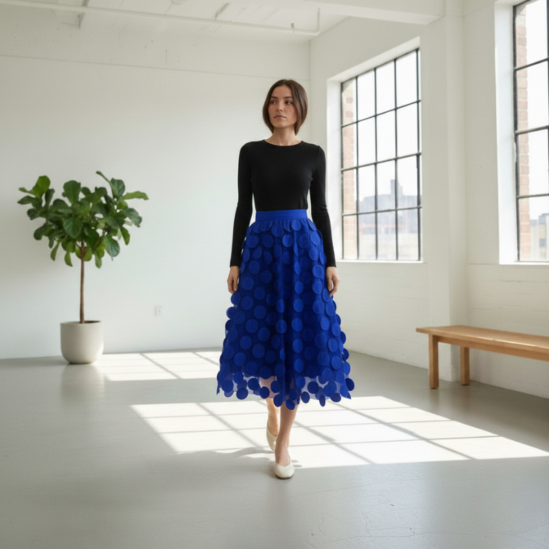 Woman wearing a black top and blue skirt in a bright room with large windows.