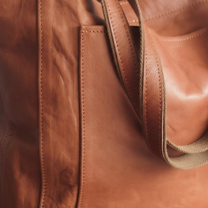 Close-up of brown leather tote bag showing stitching, grain texture, and side panel detail.