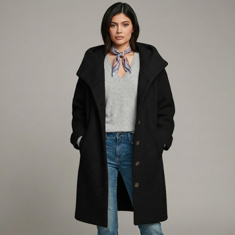 Woman wearing a black coat, gray sweater, and blue jeans on a plain background