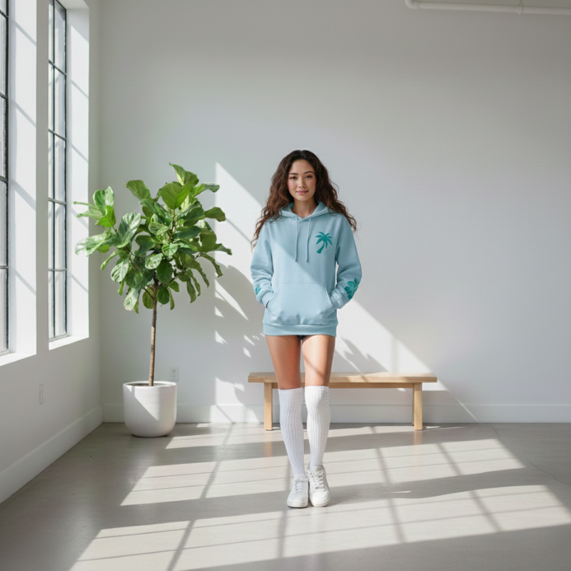 Woman in a light blue hoodie standing in a bright room with a plant and bench.